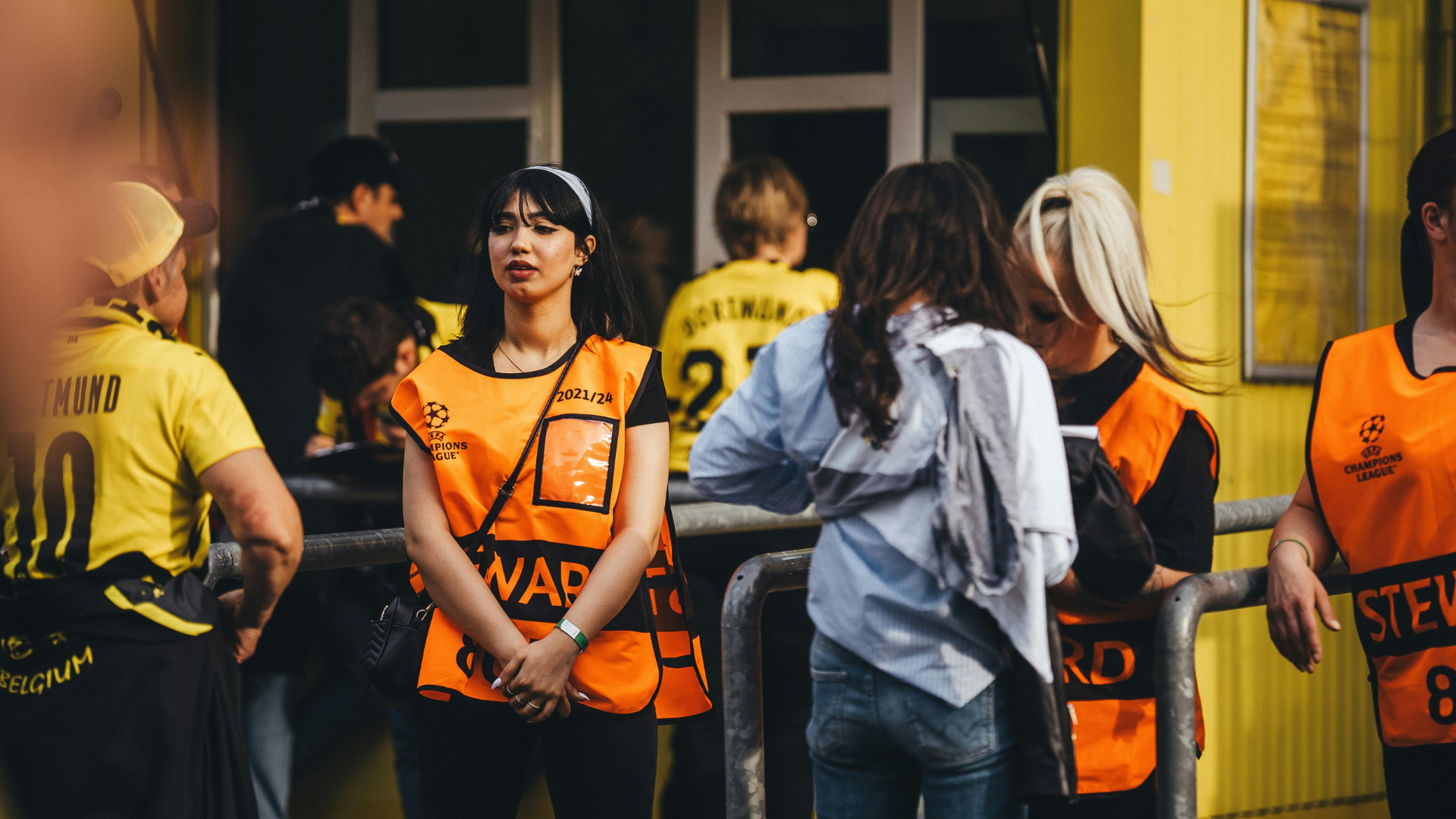 2 female stewards stood outside of a venue, 1 looking into the contents of a female patron's bag